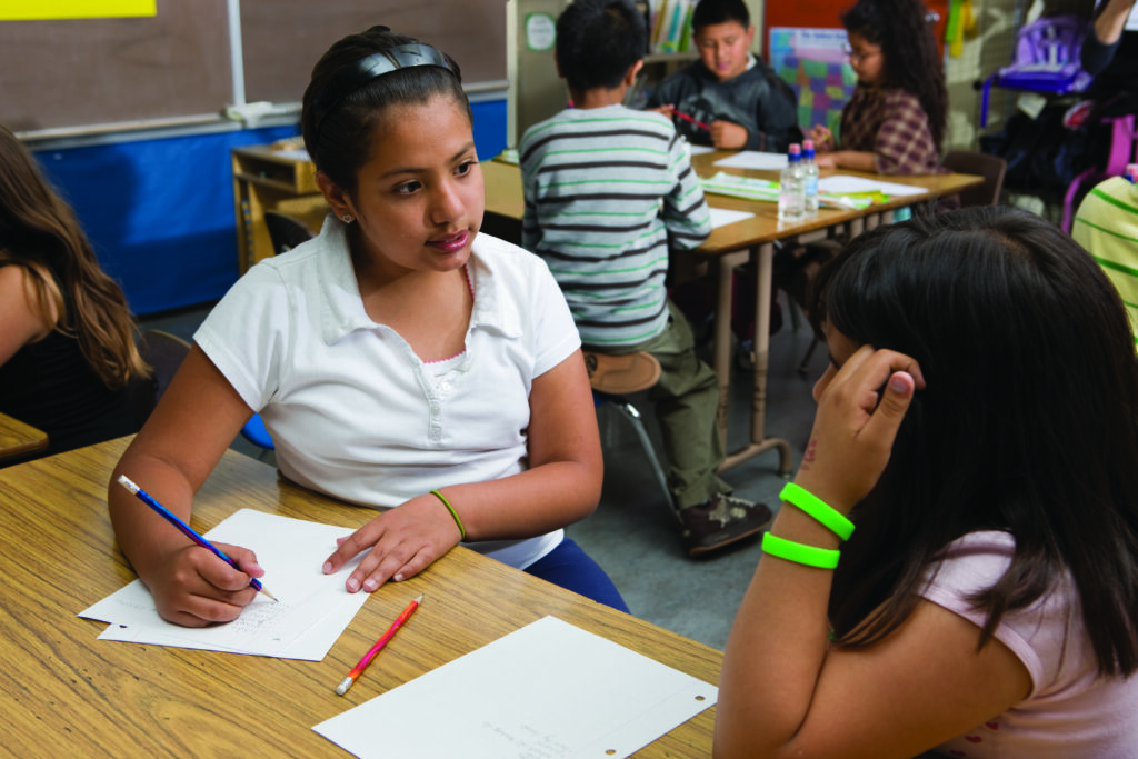 An elementary classroom where students are in groups. The focus is on two female students who discussing their writing.