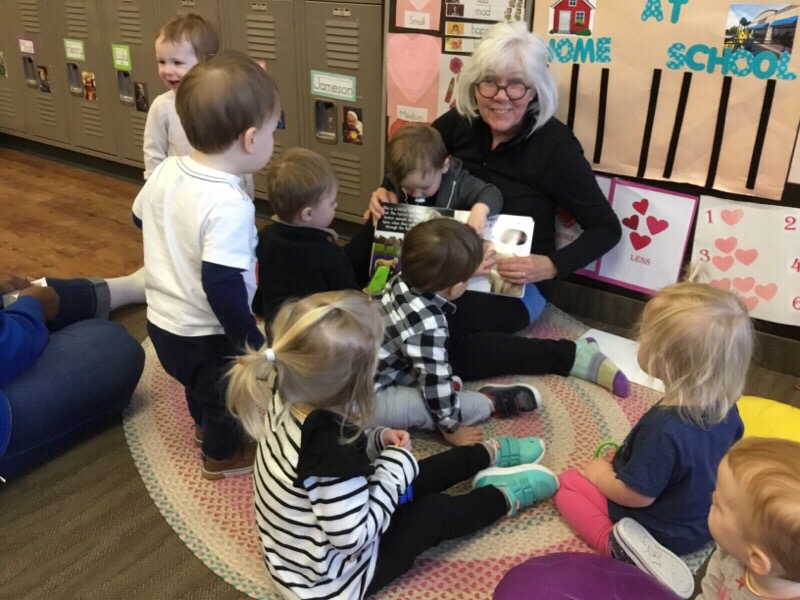 Kate Horst in a classroom sitting on a chair smiling, holding an open book in front of a group of preschoolers who are standing and sitting in front of her, pointing to pictures in the book.