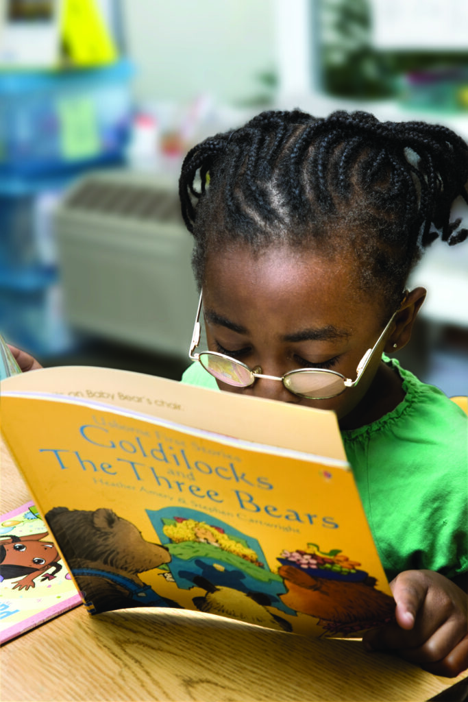 A young black female wearing glasses is holding the book "Goldilocks and The Three Bears" and reading closely.