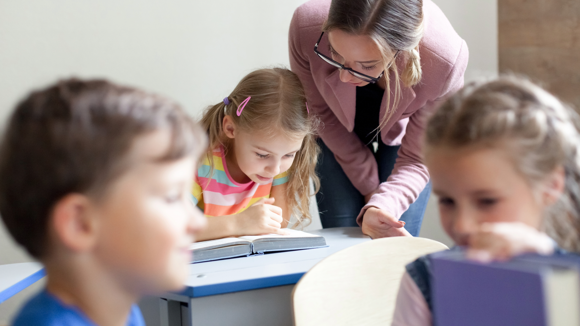 A child sits at her desk reading from a book and following along with her finger. A teacher leans over the desk watching her progress.