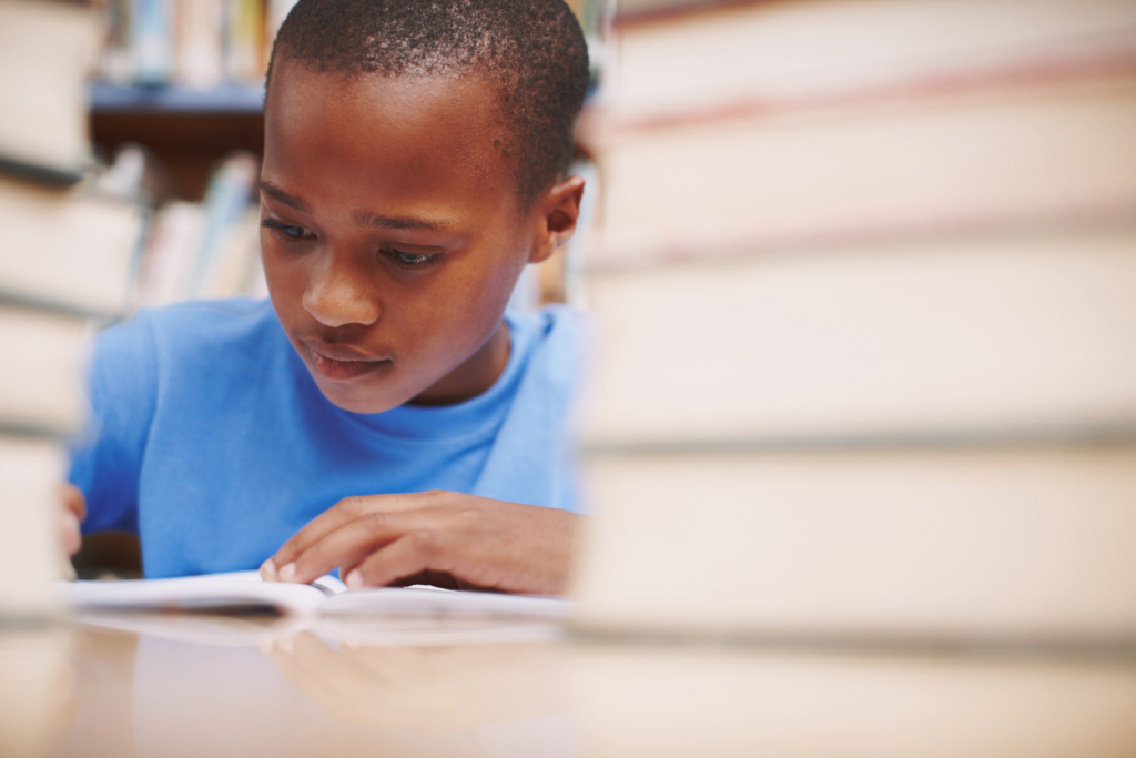 An elementary-aged child is seated at a desk surrounded by stacks of books. He holds a pen in one hand while the other hand is holding down the pages of a notebook. His eyes appear concentrated on the page in front of him.