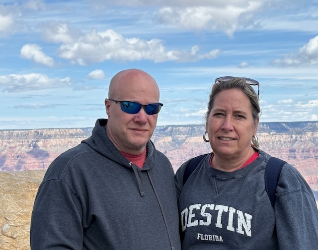 A photo of a bald man wearing sunglasses and a grey hoodie and standing next to a blonde woman with her hair back and wearing a grey Destin Florida sweatshirt. They appear to be posed in front of a canyon. This is Valerie Michaels and her husband.