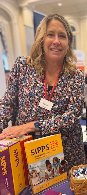 A blonde woman wearing a floral pattern top is standing next to SIPPS curriculum books. This is a picture of Valerie Michaels, A Manager of Educational Partnership for Collaborative Classroom in New York State. 