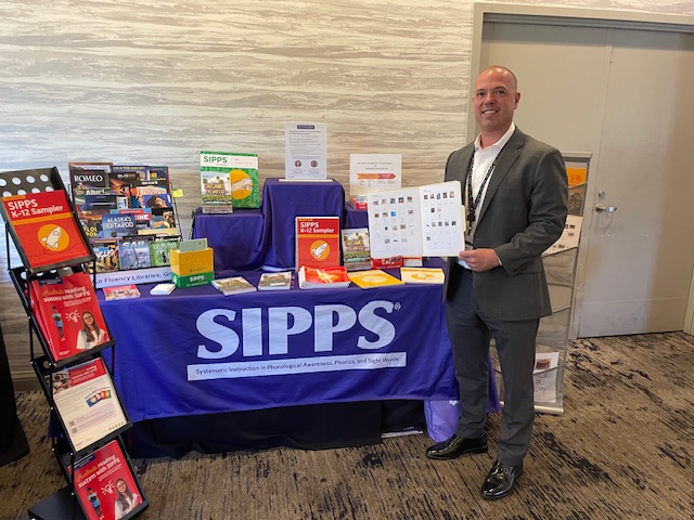 Dr. Michael Massa in front of Collaborative Classroom's SIPPS booth at the Juvenile Court, Community and Alternative School Administrators of California (JCCASAC) conference. Dr. Massa is holding a Photo Sound Spelling Card.