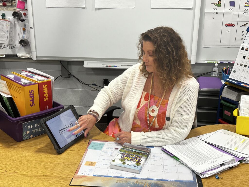 Photo shows Title 1 Reading Specialist Pamela Hunter sitting at her desk in Joe P. Eblen Intermediate School in Buncombe County, NC. Pamela is pointing at her iPad on the desk in front of her, surrounded by the SIPPS materials –including Dreams on Wheels, that she uses to meet the needs of her older readers.