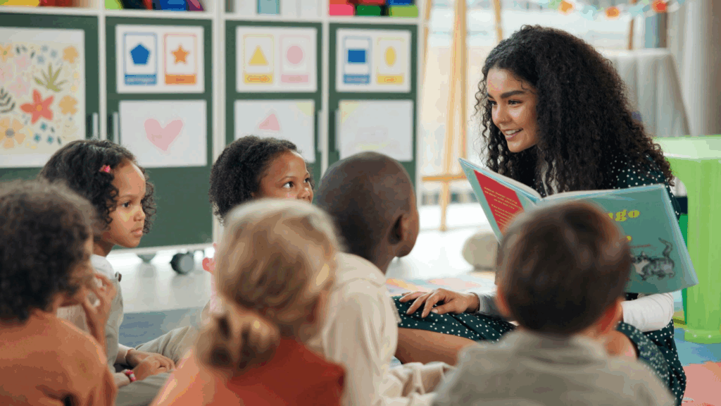 A teacher sits on the floor facing a classroom of preschool students while engaging in a repeated read-aloud. She is smiling as she looks at them, holding an open book.