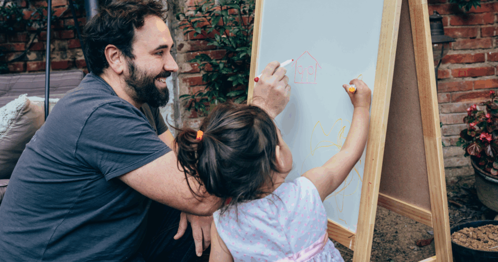 Shared writing experiences are related to the repeated read-aloud. In this photo, a teacher smiles as he shares writing on a white board with a young preschool student.