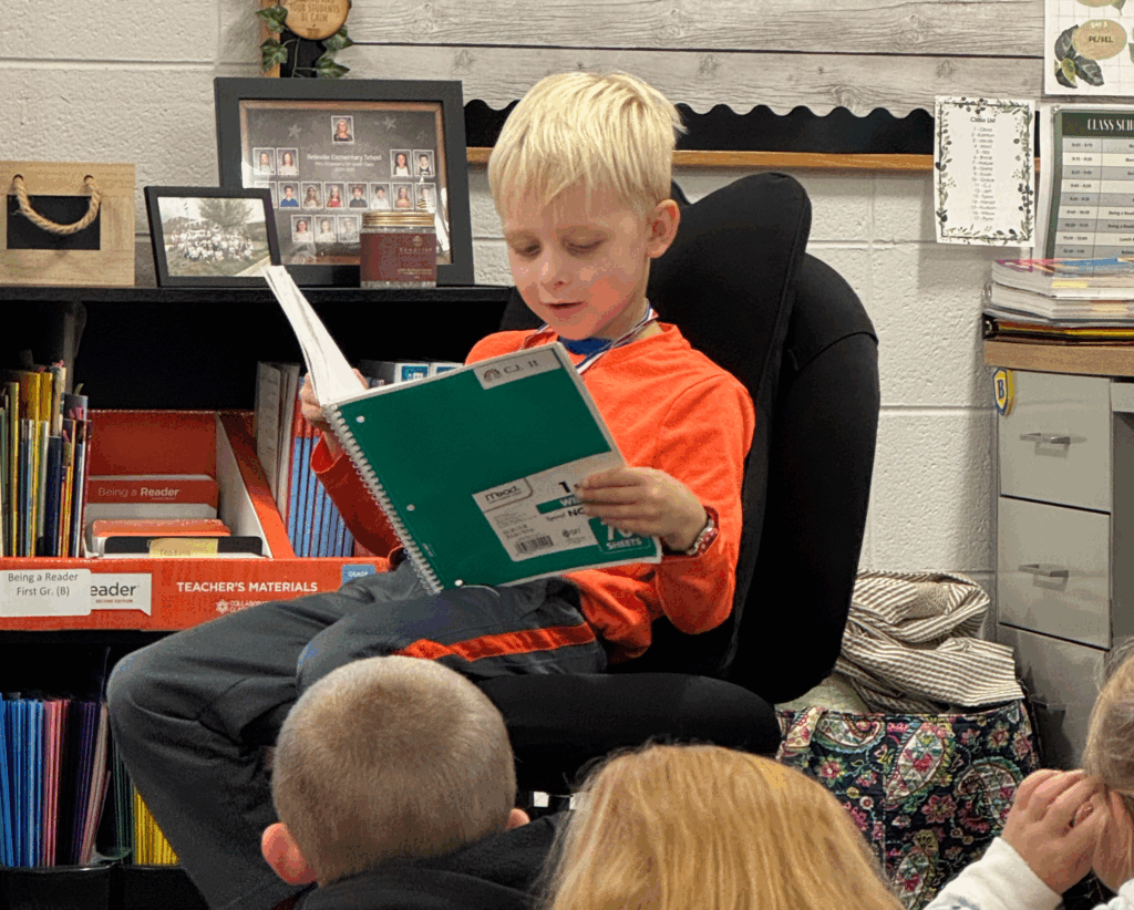 A young child sits in a chair in front of a classroom of peers, reading a book.