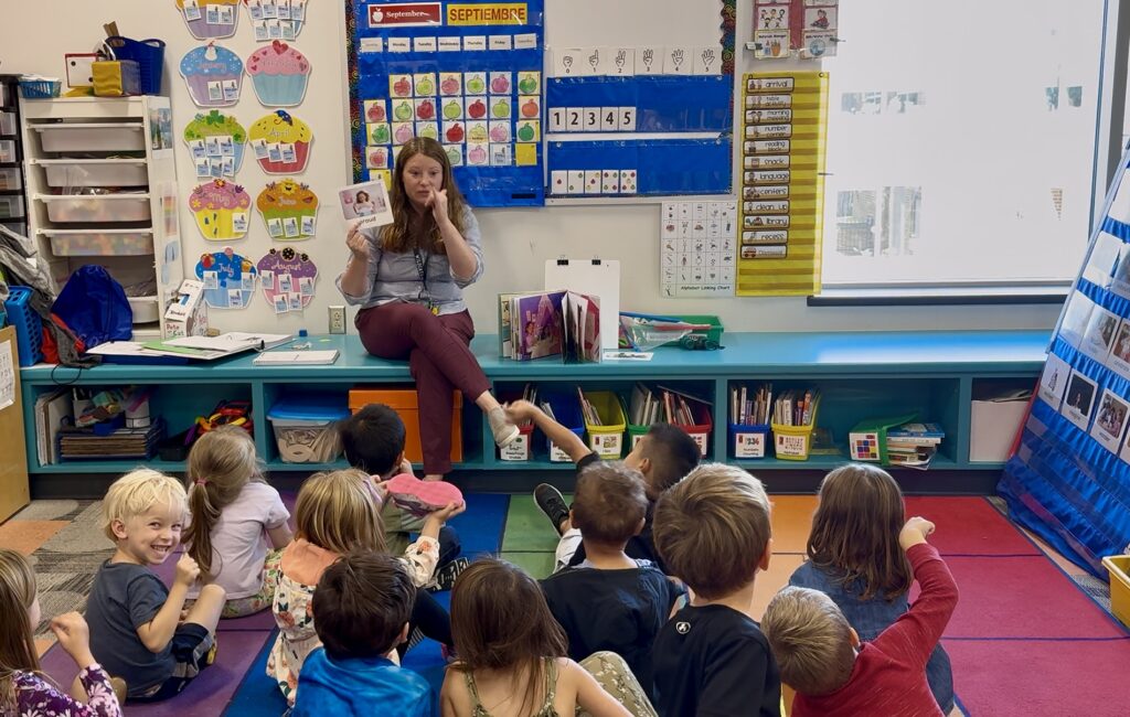 Belleville pre-k teacher sits in front of a classroom of students sitting on the floor facing her while she reads them a book.