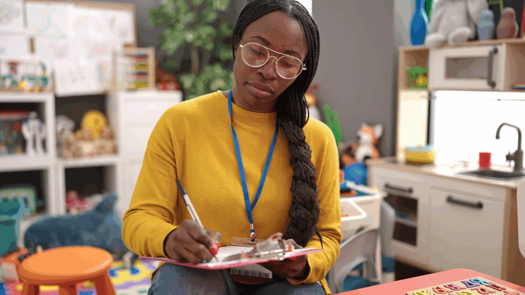 Image shows an educator wearing a yellow shirt with a lanyard around their nect holding a writing instrument and making notes on a clipboard.
