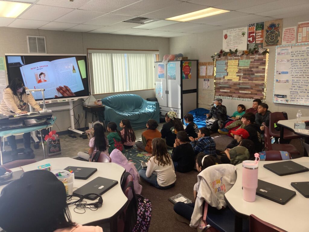 Children sit on the floor looking at and listening to a book that is projected on a screen in front of them