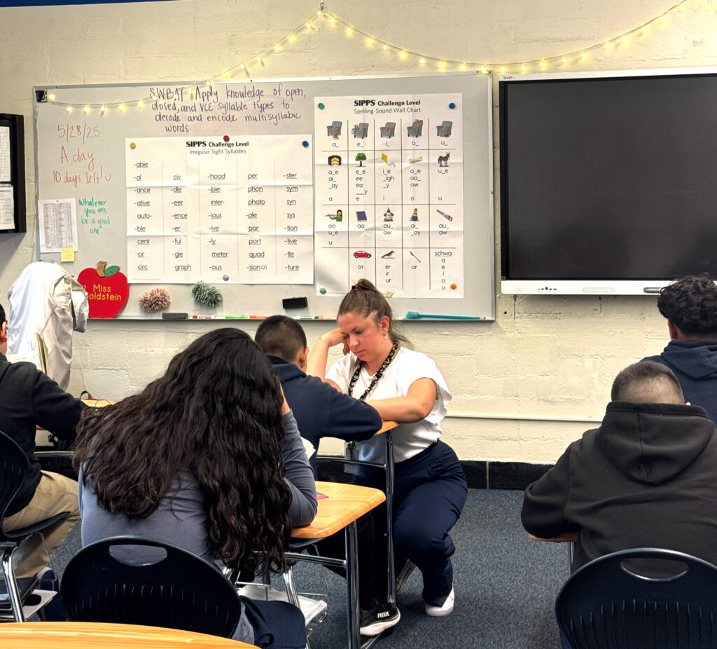 Photo shows Eisenhower Middle School teacher Molly Goldstein in a classroom, crouching next to a student who is seated at their desk.
