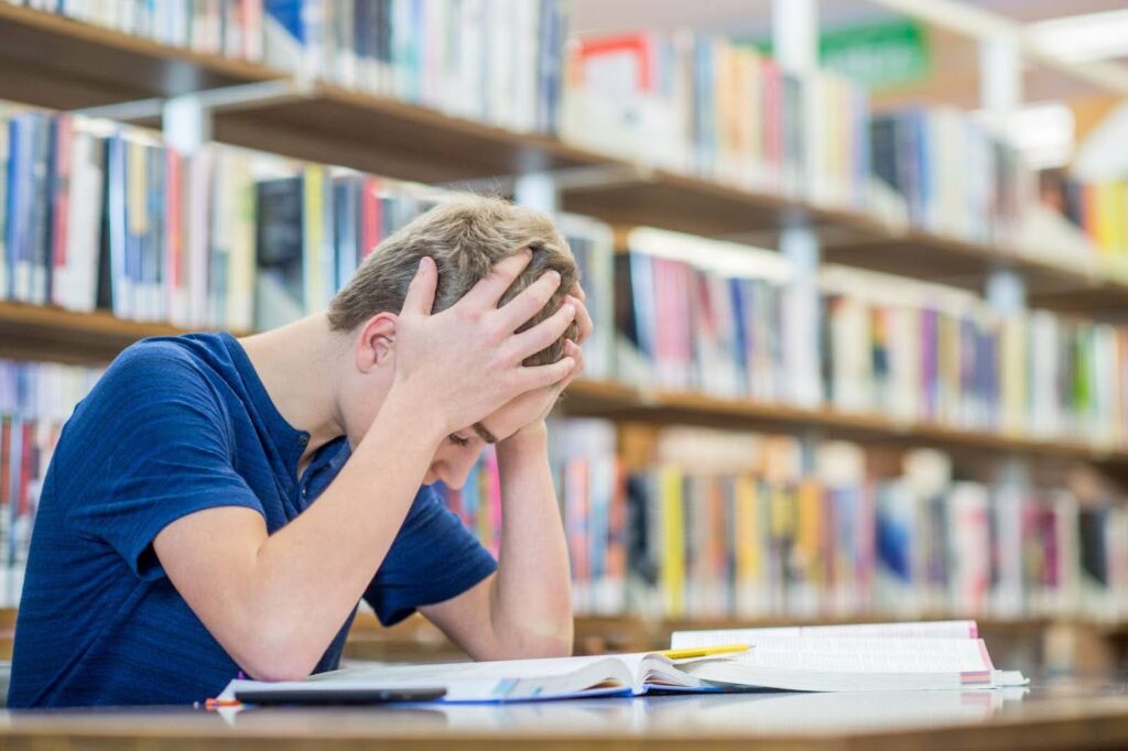 Student is sitting in a library, looking down at at a desk full of textbooks while holding their head in their hands.