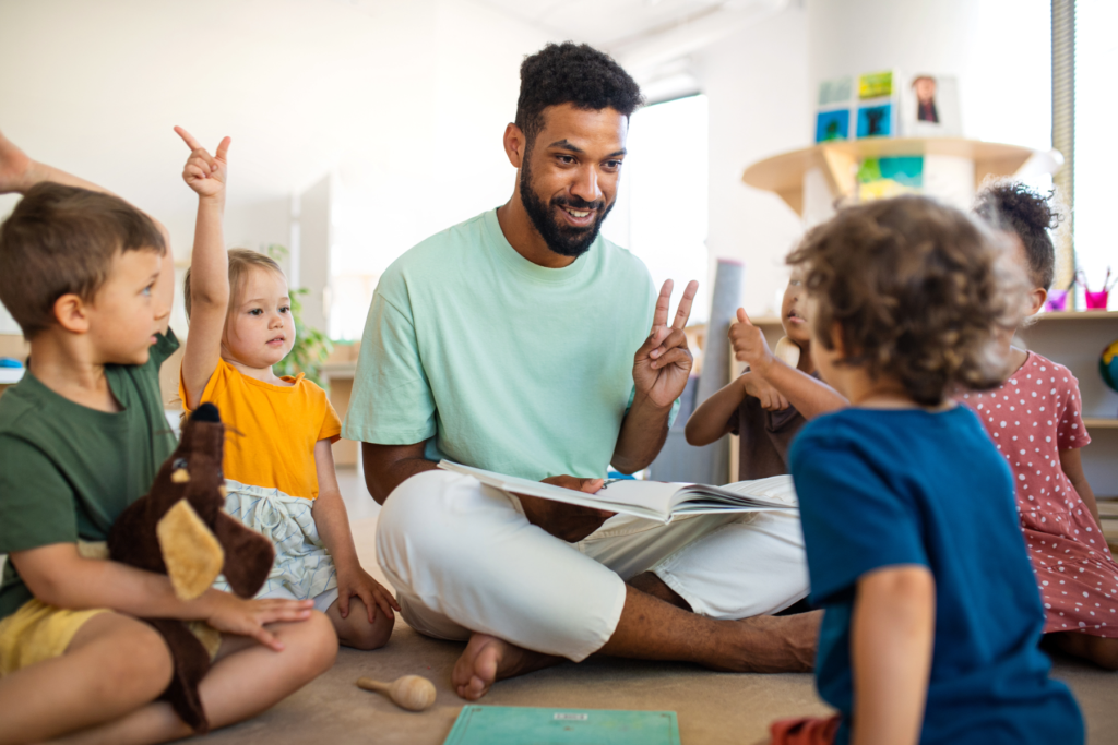 Preschool teacher does an activity with a focus on phonological awareness as he sits on the floor surrounded by children with a book on his lap and holding up two fingers.