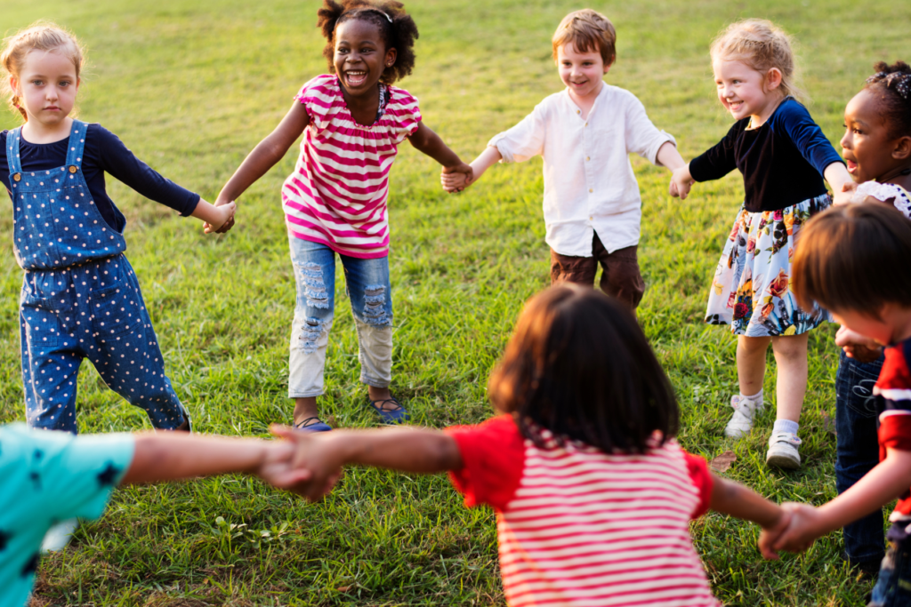 8 or more preschool children are outside holding hands in a circle playing a game focused on phonological awareness.