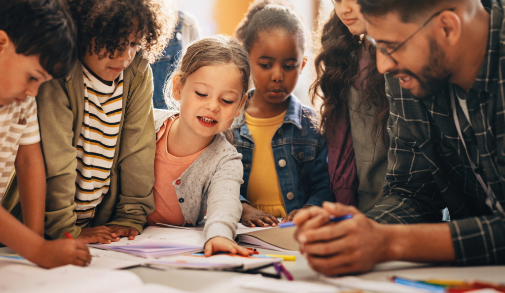 Photo shows an instructor leaning against a desk with a group of 5 children also leaning in to reach and look at books and writing utensils on the desk surface. Literacy success depends upon educators moving beyond the science of reading and embracing the three sciences framework.