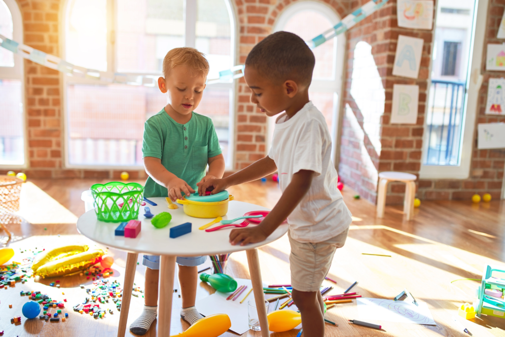Two children stand at a small table in a preschool room, talking while playing together with toys.