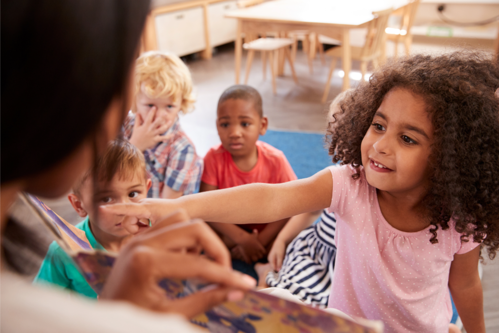 In a preschool classroom, a small child points to content in a book that the teacher is holding up in front of her and other classmates.