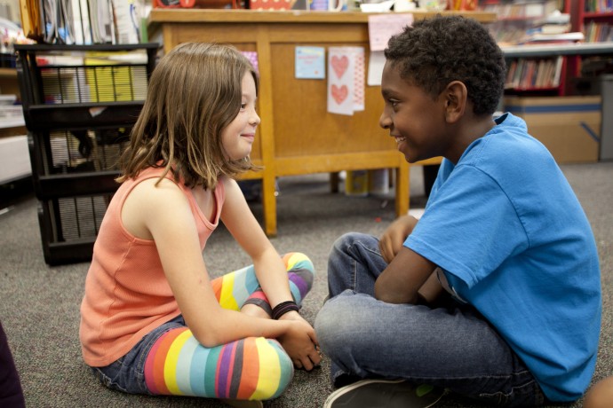 two students, facing each other and talking while seated cross-legged on a classroom floor